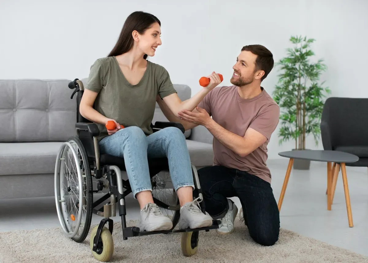 A support worker handing a dumbbell to a smiling woman in a wheelchair at home